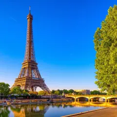Eiffeltornet i Paris, med sina tre våningar, speglar sig i Seine. En grönskande trädtopp skymtar till höger.