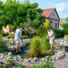Dwóch mężczyzn pielęgnuje oczko wodne w ogrodzie. Jeden czyści wodę myjką ciśnieniową, drugi wyciąga rośliny.