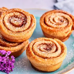 Zimtschnecken Muffins, bestäubt mit Puderzucker, liegen auf einem blauen Teller neben lila Blumen.