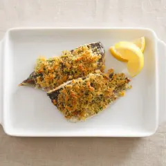 Two fillets of aji fish, baked with a golden breadcrumb topping, served with lemon wedges on a white dish.