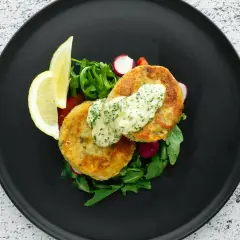 Golden chikuwa tempura patties served with a creamy herb sauce, fresh greens, radishes, and lemon wedges on a black plate.