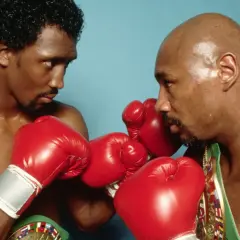 Two boxers, one with curly hair, the other bald, face off in a tense stare-down, red gloves raised, hinting at the legendary Marvin Hagler war.