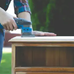 Une personne utilise une ponceuse pour lisser le dessus d'une petite commode en bois, un travail qui demande de poncer endroit difficile.