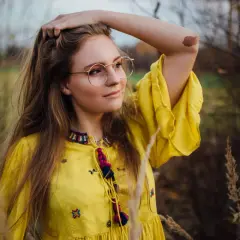 Młoda kobieta w okularach i żółtej sukience, uchwycona przez fotografa portretowego, z dłonią we włosach, na tle jesiennej przyrody.