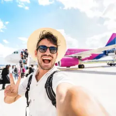 Mężczyzna w słomkowym kapeluszu i okularach przeciwsłonecznych robi selfie na tle samolotu i pasażerów.