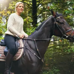 Kobieta na koniu podczas lekcji jazdy konnej w lesie. Słońce przebija się przez drzewa.