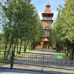 Drewniana wieża kościelna w Długosiodle, otoczona drzewami, za żelazną bramą.
