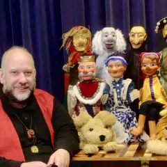 Ein Mann lächelt neben einer bunten Truppe von Handpuppen, die für den Puppenpavillon Bensberg bereitstehen.