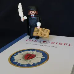 Playmobil-Figur mit Federkiel und Büchern, die den Unterschied zwischen Lutherbibel und katholischer Bibel symbolisiert.