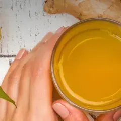 Hände halten ein Glas mit leuchtend gelbem Getränk. Daneben liegen eine Orange und Ingwer. Perfekt, um Kurkuma zuzubereiten.