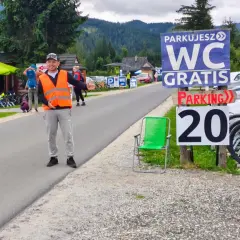 Pracownik w kamizelce odblaskowej kieruje ruchem na parkingu siwa polana. W tle góry i budynki.