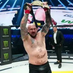 Zwycięski zawodnik KSW z pasem mistrzowskim w górze. W tle widać innych zawodników i publiczność.