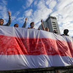 Ludzie trzymają biało-czerwoną flagę, symbol wolności Białorusi. Niebo z chmurami nad budynkiem.
