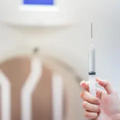Dłoń trzyma strzykawkę z płynem. W tle widać fragment aparatu do rezonansu magnetycznego, który pomaga uwidocznić szczegóły, tworząc kontrast.