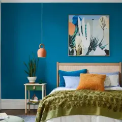 Dormitorio con **pinturas pared** azul vibrante, cama con colcha verde y cojines coloridos, cuadro abstracto y espejo ovalado.