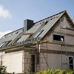 Budowa domu z silikatu w toku. Dach pokryty blachą, zamontowane okna dachowe i komin. Wokół rusztowania.