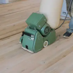 Ein Handwerker schleift mit einer Maschine einen hellen Dielenboden. Staub wird in einem Beutel gesammelt.