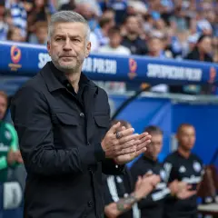 Srebrnowłosy mężczyzna w czarnej kurtce, prawdopodobnie trener roku, klaszcze na boisku.