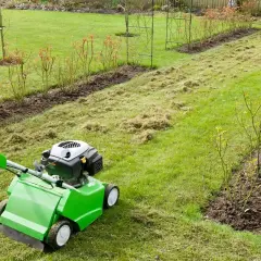 Osoba w rękawiczce obsługuje zieloną aeratorę, która wykonuje nakłuwanie trawnika, usuwając filc i mech.