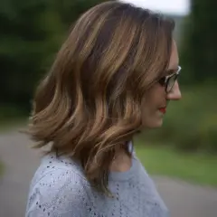 Une femme aux mèches brunes sur cheveux blancs, portant des lunettes, regarde au loin dans un décor verdoyant.