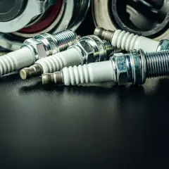 Kilka świec zapłonowych, niektóre z widocznym nalotem, leżą obok części mechanicznych.