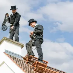Zwei Schornsteinfeger in Zylinder und traditioneller Kleidung stehen auf einem Dach. Sie bereiten sich auf ihre Arbeit vor, denn bei der Wahl des Schornsteinfegers zählt die Qualität.