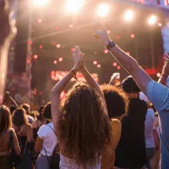 Folla entusiasta applaude e balla sotto le luci di un concerto. Per godersi al meglio l'evento, pensa a cosa portare ad un concerto: acqua, crema solare e un sorriso!