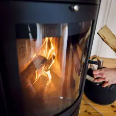 Ein Kaminofen mit brennendem Holz. Eine Hand hält ein Holzscheit, bereit zum Nachlegen. Bald muss der alte Kaminofen entsorgt werden.