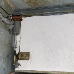 Detailaufnahme einer Brandschutzklappe mit sichtbaren mechanischen Teilen und einer weißen Platte. Möglicherweise Asbest-verdächtige Materialien.