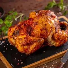 Ein goldbraunes, knuspriges Hähnchen liegt auf einem Holzbrett, bereit zum Servieren. Daneben liegen ein Messer und eine Gabel.