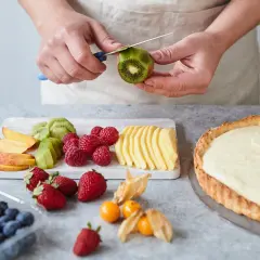 Hände schälen Kiwi für ein fruchtiges Tarte Rezept. Daneben liegen Beeren, Pfirsichspalten und Apfelscheiben.