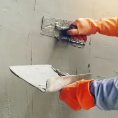 Ręce w pomarańczowych rękawicach nakładają gips na ścianę. Zastanawiasz się, ile za tynki gipsowe?