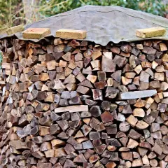Ein großer Stapel Brennholz, der mit einer Plane und Ziegeln abgedeckt ist. Die Holzscheite sind ordentlich aufgeschichtet, bereit für den Winter. Die Baustahlmatte dient als stabile Unterlage.