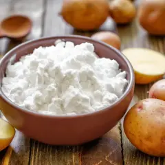 Miska pełna białego krochmalu ziemniaczanego, obok ziemniaki i drewniana łyżka. Idealne składniki do domowej kąpieli w krochmalu.