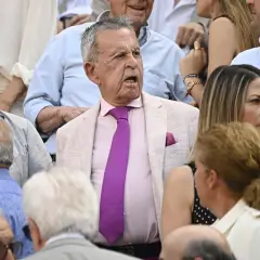 Un grupo de personas, entre ellos algunos famosos en las ventas, observan atentamente un evento. El hombre del centro, con traje rosa y corbata morada, parece sorprendido.