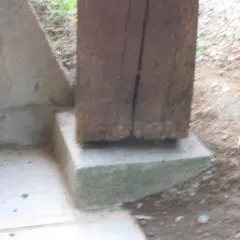 Un poteau bois pourri à la base repose sur un socle en béton. La dégradation du bois est visible.