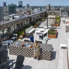 Ein begehbarer Flachdachgarten mit Stadtblick. Menschen entspannen sich auf der Dachterrasse, die mit Sitzgelegenheiten, Pflanzen und einem Schachbrett-Tisch ausgestattet ist.