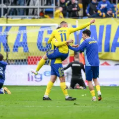 Piłkarze w żółto-niebieskich strojach świętują zwycięstwo na boisku, jeden z nich jest niesiony przez kolegę.