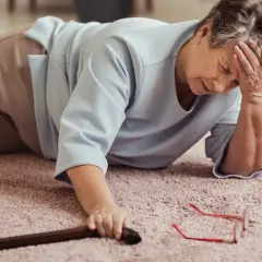 Une femme âgée gît sur le sol, la tête dans la main, ses jambes qui flageolent. Sa canne et ses lunettes sont tombées à côté d'elle.