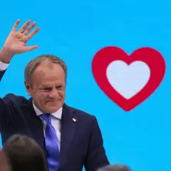 Donald Tusk macha do tłumu na tle niebieskiego ekranu z czerwono-białym sercem, symbolem Koalicji Obywatelskiej.