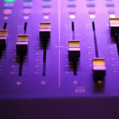 Panel miksera dźwiękowego z faderami i podświetlanymi przyciskami w fioletowym świetle.