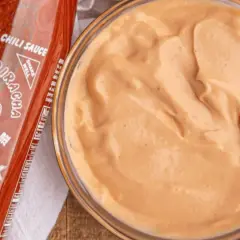 A bowl of creamy, orange spicy mayo recipe sits next to a bottle of Sriracha hot chili sauce.