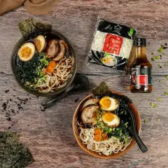 Dwa miski aromatycznego ramen z jajkiem, mięsem i wodorostami. Obok makaron ramen i sos.