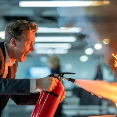 Mężczyzna w garniturze desperacko próbuje gaszenie pożarów za pomocą gaśnicy, która płonie na tablicy z planem projektu.