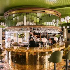 Barman préparant un cocktail dans un bar chic aux murs verts et au comptoir doré. Une ambiance de "pink mama" pour une soirée réussie.