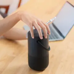 Dłoń dotyka czarnego głośnika, obok laptopa. Dowiedz się, jak podłączyć głośniki do komputera, aby cieszyć się lepszym dźwiękiem.