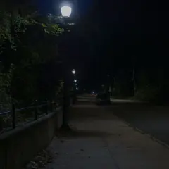 Nocna ulica z latarniami, chodnikiem i samochodami. Ciemność i światło lamp tworzą atmosferę jak z filmu, gdzie nawet kurza ślepota wydaje się możliwa.