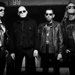 Czterech mężczyzn w skórzanych kurtkach, w tym jeden z napisem "Sisters of Mercy" na koszulce, pozuje do zdjęcia.