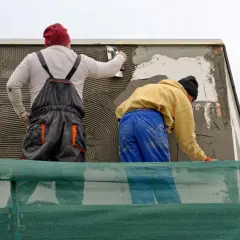 Lavoratori applicano rasatura armata con rete in fibra di vetro su un muro esterno, protetti da un telo verde.