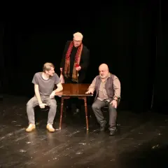 Trzej mężczyźni na scenie Teatru Korez, Plac Sejmu Śląskiego Katowice. Jeden stoi, dwóch siedzi przy stole.